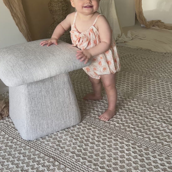 Baby wearing yellow romper standing near chair with decorative dried palm leaves