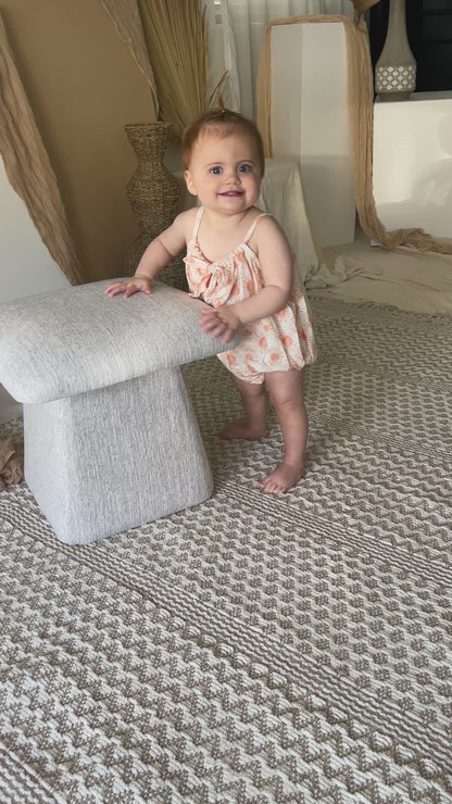 Baby wearing yellow romper standing near chair with decorative dried palm leaves